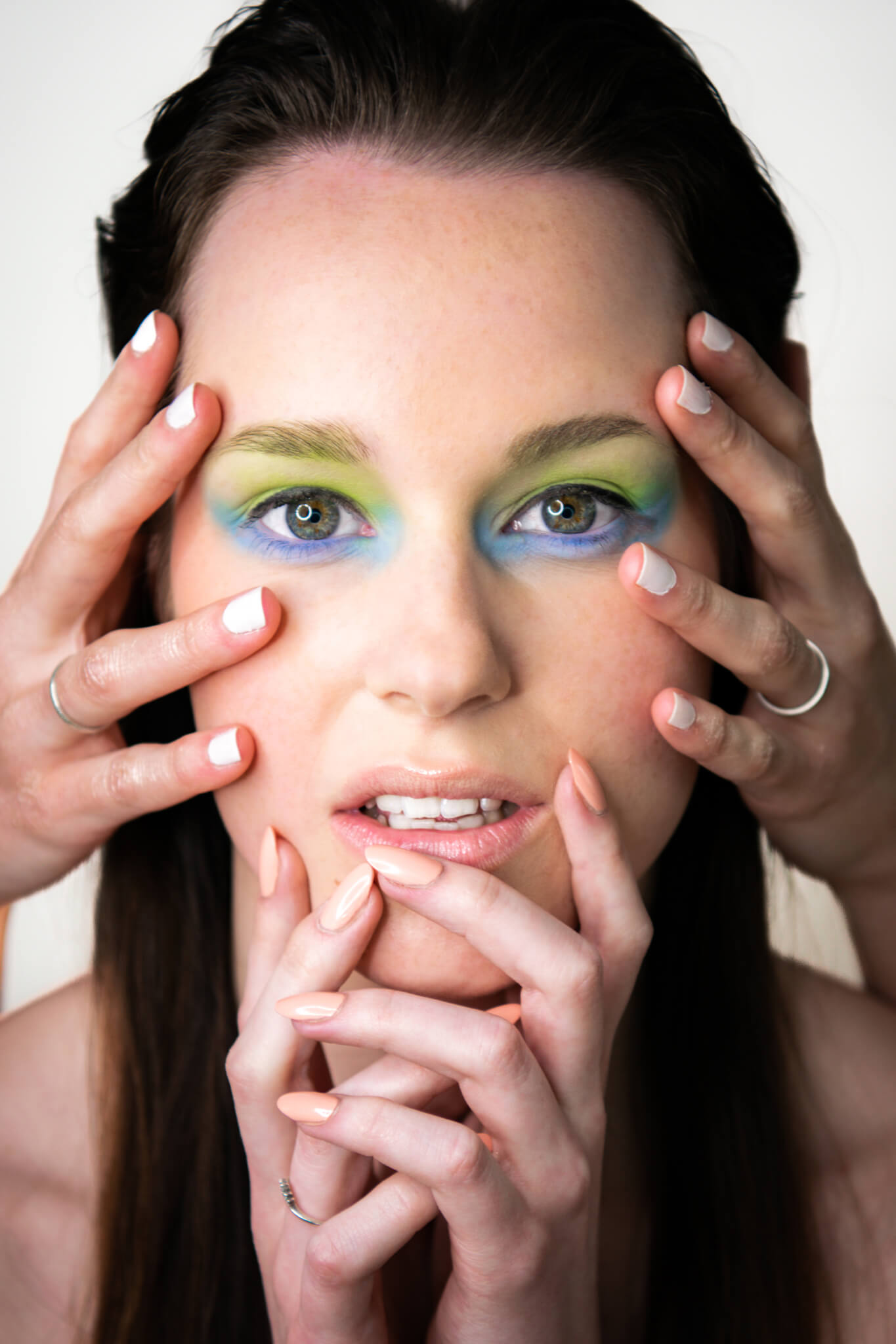 Dunkelhaarige, junge Frau mit grün-blauem Augenmakeup mit vier Händen auf ihrem Gesicht